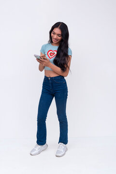 A smiling asian trans woman chatting with her boyfriend or friend on her cellphone. Wearing a light blue printed shirt and jeans. Whole body photo on a white background.