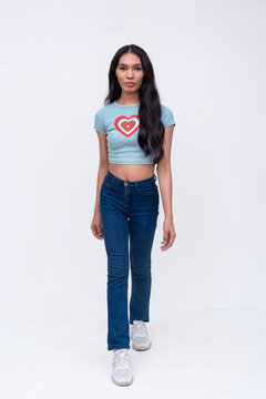 A slim trans woman walking slowly like a model. Sauntering wearing a light blue printed shirt and jeans. Whole body photo on a white background.