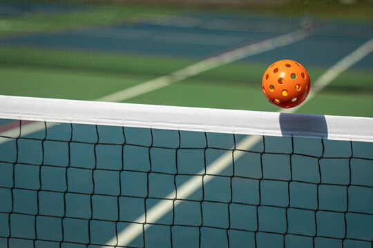 Orange Pickleball crossing a Pickleball net with shadow on the top tape.