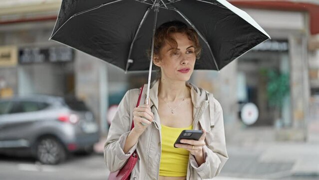Young woman holding umbrella using smartphone smiling at street