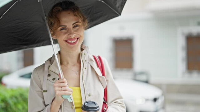 Young woman holding umbrella and coffee smiling at street