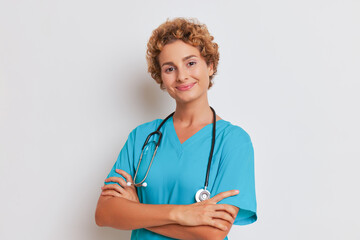 Young curly woman stands with hands crossed on her chest, beautiful female doctor smiling gently on white background, good doctor concept, copy space