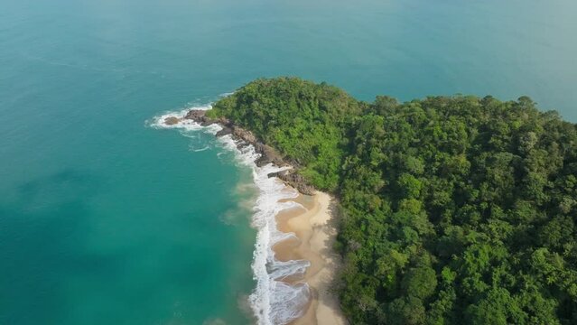 A drone descends, approaching some rocks with waves crashing, surrounded by trees and foamy white waves. The horizon reveals a vast blue ocean and beaches in the distance. 4k resolution, Brazil, parad