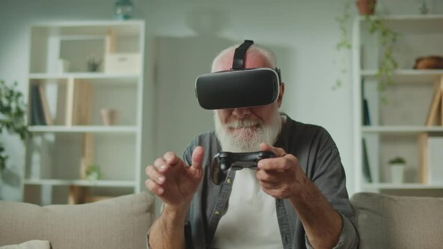 An Old Man in VR Glasses Plays Computer Games With a Joystick, Sitting in the Sofa and Eating a Popcorn.A Modern Elderly Man Plays PlayStation. Augmented Reality and Gaming for Seniors.