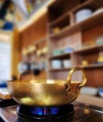 Brass wok on gas stove with flame and blurred kitchen atmosphere as background.