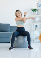 Asian young sexy pretty fit female athlete teenager in sportswear sport bra leggings and watch sitting smiling stretching arms and hands on exercise ball warming up working out in living room at home