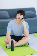 Asian young handsome fit male athlete teenager in sportswear sneakers sitting smiling on yoga pilates mat taking coffee break holding water bottle and smartphone in hand on call in gym after training