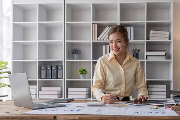 Beautiful and happy young Asian businesswoman looking at her laptop screen while enjoying her morning coffee at her desk in the office.