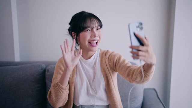 Happy young asian woman having video call and waving hand to friend on mobile cell phone or smartphone in living room relaxing at home.