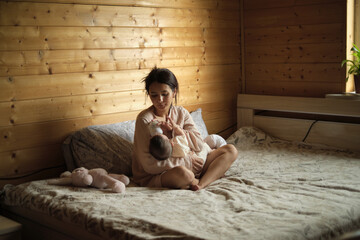 Sitting on the bed, a young mother feeds her baby with a specialized formula, demonstrating the diverse choices parents make today, sometimes opting for or relying on alternative feeding methods.