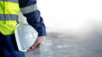 Safety workwear concept. Male hand holding white safety helmet or hard hat. Construction worker man in protective suit and reflective green vest standing with building concrete floor in the background