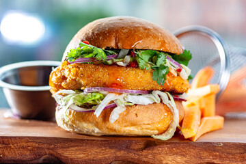 Hamburger with chicken cutlet, fresh tomatoes, red onion, lettuce and tomato sauce. Next to the burger is a metal mold with fries, a metal mold with chili sauce. Burger, fries and sauce are on a brown