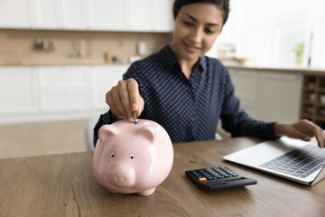 Obraz na płótnie Canvas Positive young Indian freelance business woman saving money for future, dropping coin into pink piggy bank on workplace table at home, enjoying financial management, investment