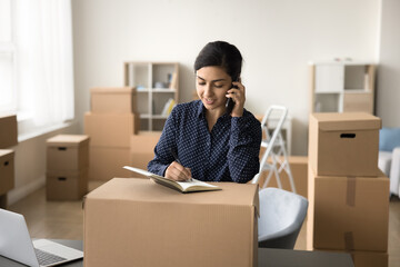 Pretty young Indian storage manager girl sending purchase from Internet store to customer, talking on mobile phone at packed cardboard box, writing notes, processing order