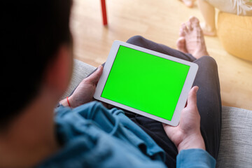 Over the shoulder view of a man working on a tablet with a green chroma key screen while sitting on a couch