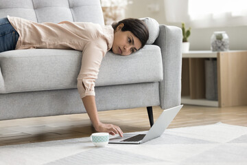 Overworked exhausted Indian freelancer woman lying on sofa with face on seat, using laptop computer...