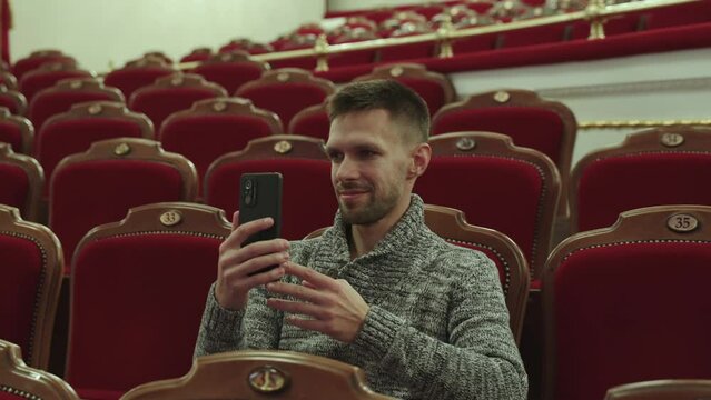 Hispanic middle aged man takes pictures of auditorium in theater on phone. Brunet in sweater sits in comfortable burgundy velvet chair and waits for show to begin, handheld shot.