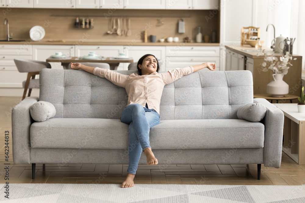 Wall mural cheerful positive indian new homeowner stretching body on stylish comfortable couch in cozy home int