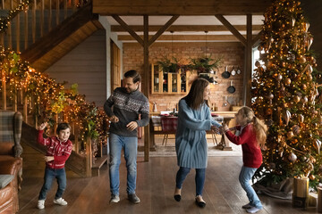Fototapeta na wymiar Happy young couple parents dancing to energetic disco music with adorable little children son daughter near Christmas tree in beautifully decorated house, New Year holidays celebration concept.