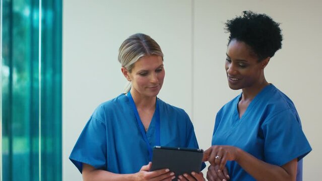 Two smiling mature female doctors wearing scrubs meeting and checking digital tablet in hospital - shot in slow motion