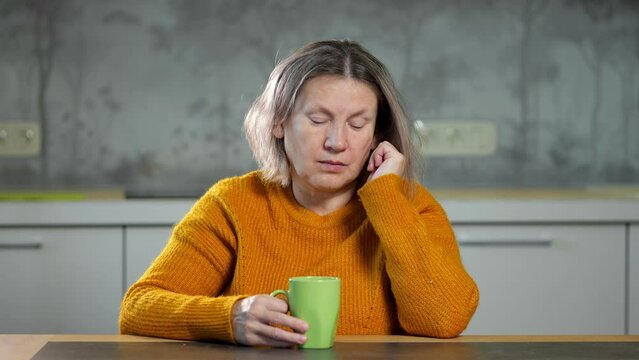 Senior woman yawns and naps during breakfast in apartment. Woman with short frizzy hair drinking hot coffee from cup to energize for day