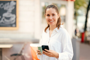 Young woman enjoys her coffee while holding phone and smiling at the camera.