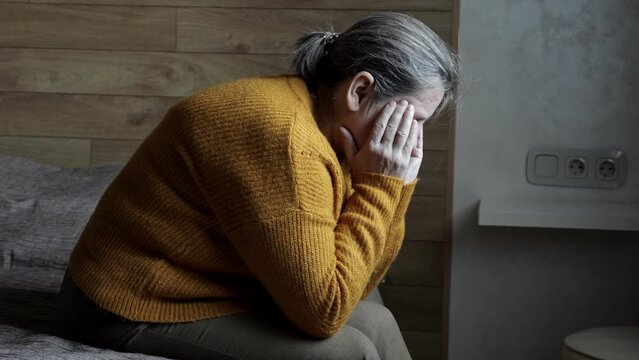 Mature woman with severe headache sitting on regular bed in bedroom. Mature woman touching head with wrinkled hands in semi-dark bedroom