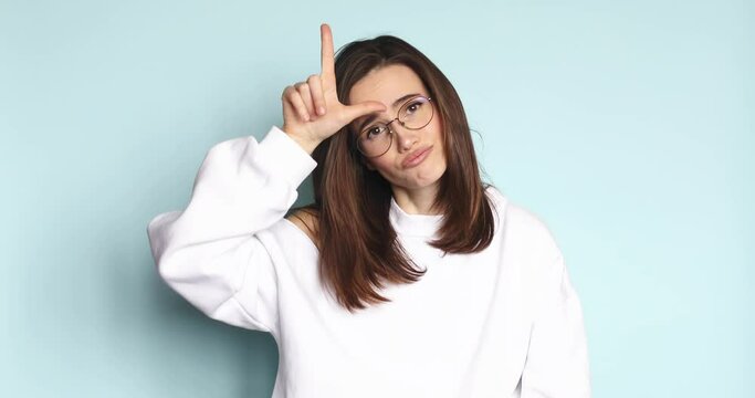 You lose! Brunette young woman making loser gesture, L sign on forehead, teasing and accusing for defeat, expressing disrespect. Indoor studio shot isolated on blue background.