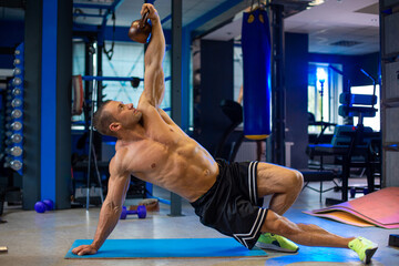 Athletic fit sporty muscular caucasian man pushes brown iron kettlebell in contemporary gym topless in black shorts and lime green sneakers, blurred bluish background