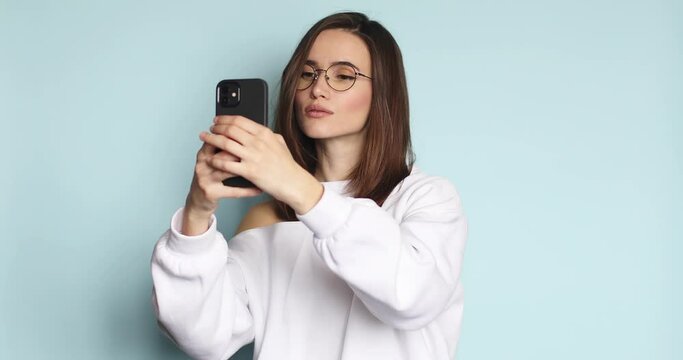 Young girl in eyeglasses isolated over blue background take a selfie looking at phone as in a mirror. Girl look calm and sexy. Short haircut.