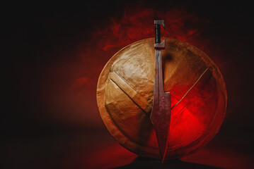 Fototapeta na wymiar Ancient Spartan sword and shield on the dark background in the red light smoke concept. Spartan concept background with copy space.