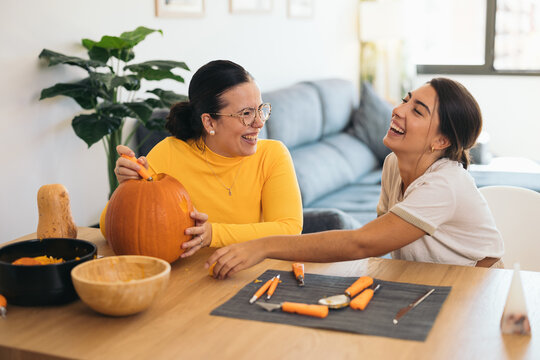 Happy young ethnic women sitting at table with orange pumpkin and carving eye