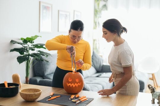 Focused young ethnic women standing and putting candle into pumpkin