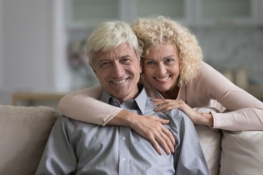 Cheerful happy senior retired married couple home portrait. Pretty wife hugging grey haired husband, looking at camera with toothy smile, enjoying good close love relationships, marriage, romance