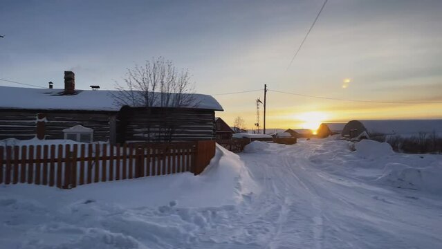 View from a car passing a wooden house. Old village building. Beautiful winter sunset. Large layer of snow on the road.