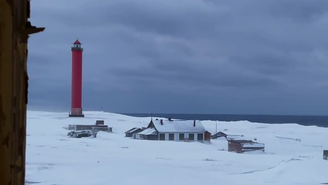 Big red lighthouse in a winter village. Wooden houses in the wasteland. snow covered field. Dark sky with clouds