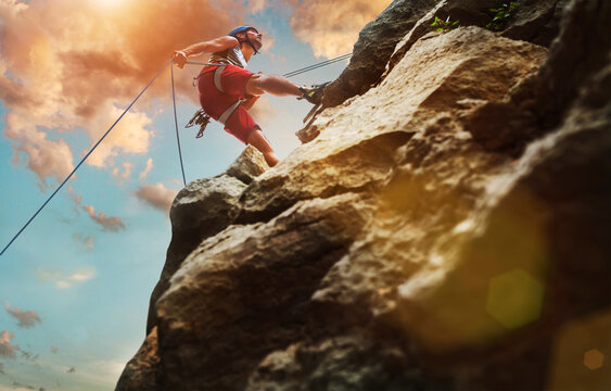 Muscular climber man in protective helmet abseiling from cliff rock wall using rope Belay device and climbing harness on evening sunset sky background. Active extreme sports time spending concept.