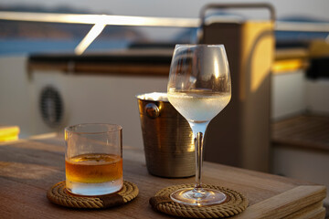 glass of white wine and whiskey as lady and gentleman cold beverages on yacht sunset sail served on teak wood natural table