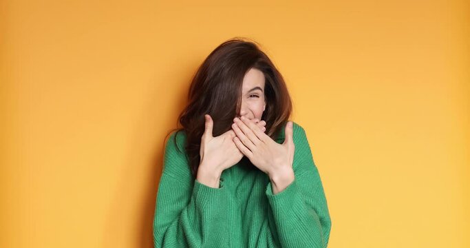 Shocked shy charming confused young woman in green sweater posing isolated on yellow background studio. Girl looking at camera smile cover mouth with hands say oops.