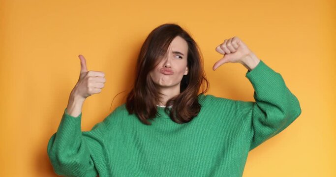 Young woman in green sweater making good-bad sign isolated on yellow background. Girl make choice, or makes a decision thumb up or tnumb down, like or dislike, yes or no. 