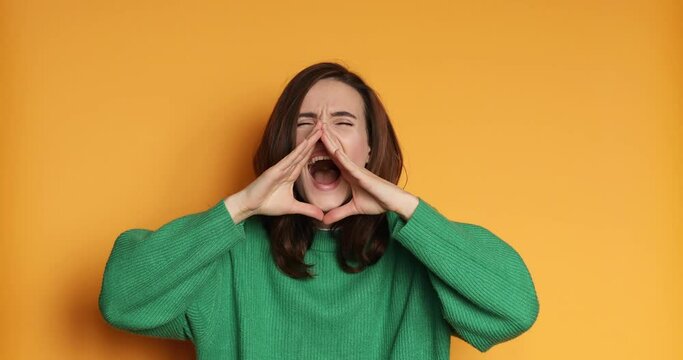 Angry woman wear green sweater screaming on yellow background. Portrait of angry girl scream in studio. Rage woman screaming and hold hands near face. Aggressive person shouting in studio