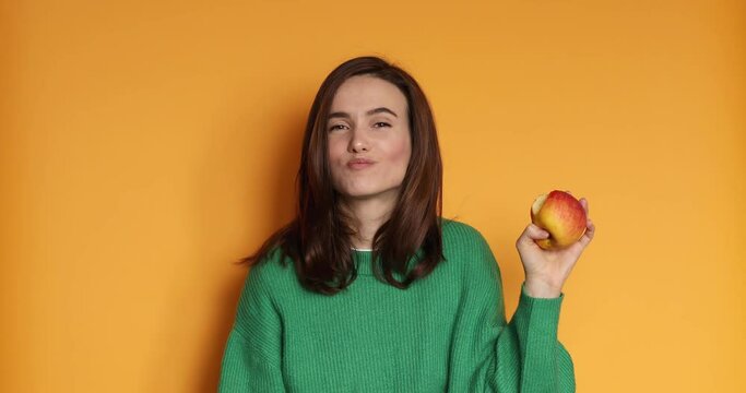 Young brunette woman hold apple, eating apple and smile. Bite fresh apple with happiness to diet, weight and calories control and good health concept isolated on yellow background. Healthy teeth.
