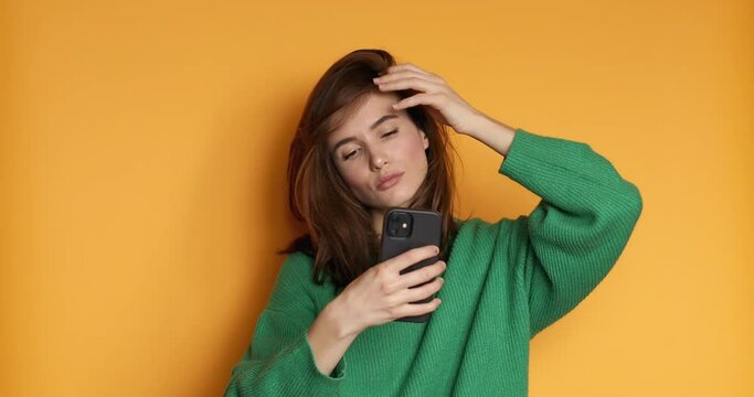 Brunette girl stares at the phone and fixes her hair. Female uses phone like mirror. Young girl make photo from hands with phone on yellow background.