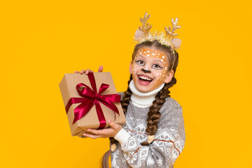 A happy little girl holds a gift box with a red bow. A child with a carnival make-up of a deer and horns is enjoying the holiday. Merry Christmas. Bright yellow isolated background.