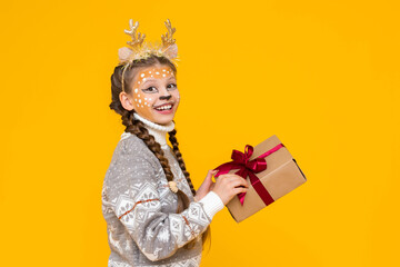 A little girl is dressed in a carnival festive deer with horns. Child holds a gift box with a red bow. New Year's holidays for children. Merry Christmas. Yellow isolated background.