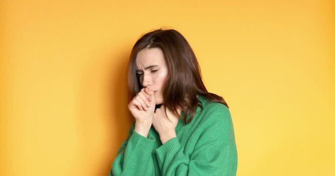 Woman wear green sweater is suffering with cough and feeling bad on yellow background. Health care and safety. Millennial woman coughing hardly.