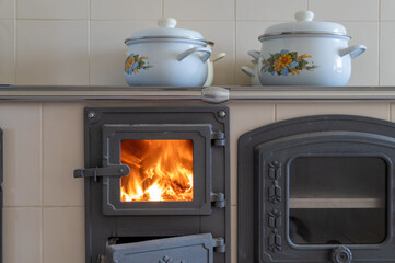 Wood burning tiled stove with fire in the kitchen with white pots on top.
