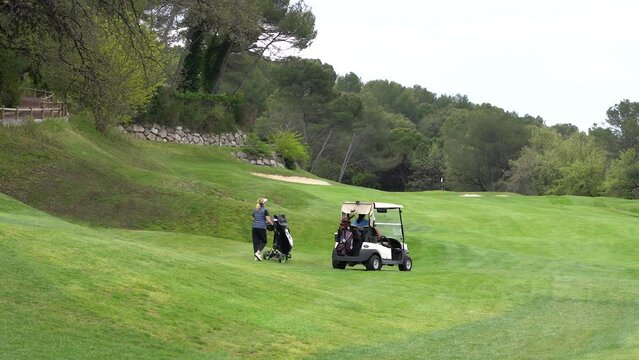A woman walks along a golf course, carrying a cart for clubs in front of her, and a car for transporting golfers and equipment drives nearby.
