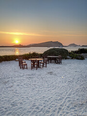 Two wooden tables with chairs on a white sandy beach by the ocean at sunset and islands and coastline visible on the horizon, taken on the island of St Martins, Isles of Scilly