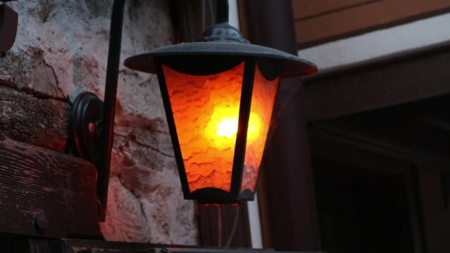 Metal lantern with an old design and a flame inside hangs on a medieval stone wall. It casts a warm orange light and creates a contrast with the dark background in a vintage and rustic style
